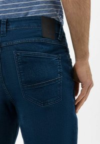 Jean en denim bleu foncé avec une coupe droite, poche arrière visible et un patch en cuir noir à la taille. La texture lisse renforce la durabilité.