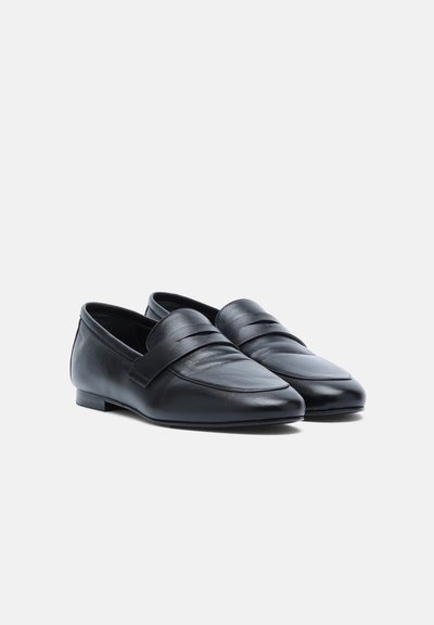 Paire de mocassins noirs en cuir à enfiler avec des talons bas et une bride décorative sur le dessus, sur un fond clair uni.