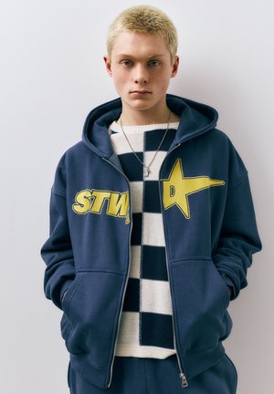 Młoda osoba o krótkich blond włosach nosi granatową bluzę na zamek z żółtym nadrukiem, czarno-białym kraciastym sweterkiem oraz srebrny naszyjnik, ma ręce w kieszeniach.