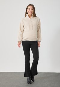 Beige fliisist pullover, millel on pooltõmbluku krae, tekstuuriga pind ja elastsed varrukad, millele on lisatud mustad flöödid leggingud ja pahkluu saapad.