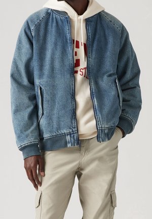 Homme portant une veste bomber en denim bleu sur un sweat à capuche beige avec un texte rouge et un pantalon cargo beige, debout avec une main dans la poche.