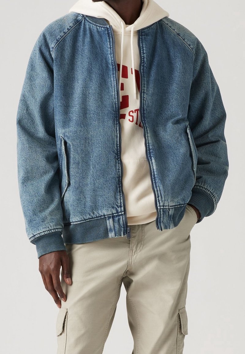 Homme portant une veste bomber en denim bleu sur un sweat à capuche beige avec un texte rouge et un pantalon cargo beige, debout avec une main dans la poche.
