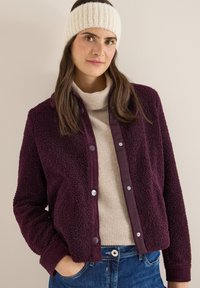 Bordowa kurtka sherpa z prążkowanym kołnierzem i zatrzaskami. Nosi się ją na beżowym golfie i łączy z denimem. Miękka faktura, casualowy design.