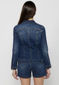 Temno modra denim jakna s prilegajočim krojem, vidnimi šivi in gumbi. Ob šortih iz denima, ki prikazujejo podrobnosti in teksturo hrbta.
