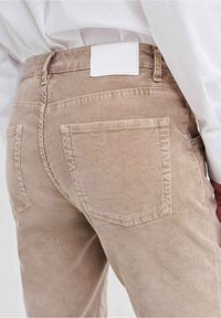 Close-up of beige corduroy pants worn with a white shirt, showing back pockets and a blank white label on the waistband.
