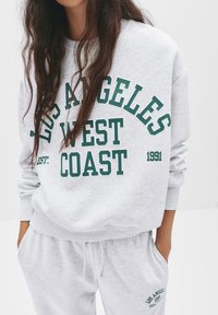 Grijze katoenen sweatshirt met groene opdruktekst "LOS ANGELES WEST COAST," met een ronde halslijn en ribgebreide manchetten. Gecombineerd met bijpassende joggingbroek.