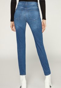 Jeans de mezclilla azul ajustados con una cintura de altura media, cinco bolsillos y una textura suave. La parte trasera presenta un mínimo detalle de costura.