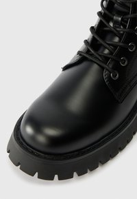 Černé kožené kotníkové boty s kulatou špičkou, texturovanou podrážkou a vyvýšeným švem. Obsahují černé tkaničky a kovové očka. Robustní design.