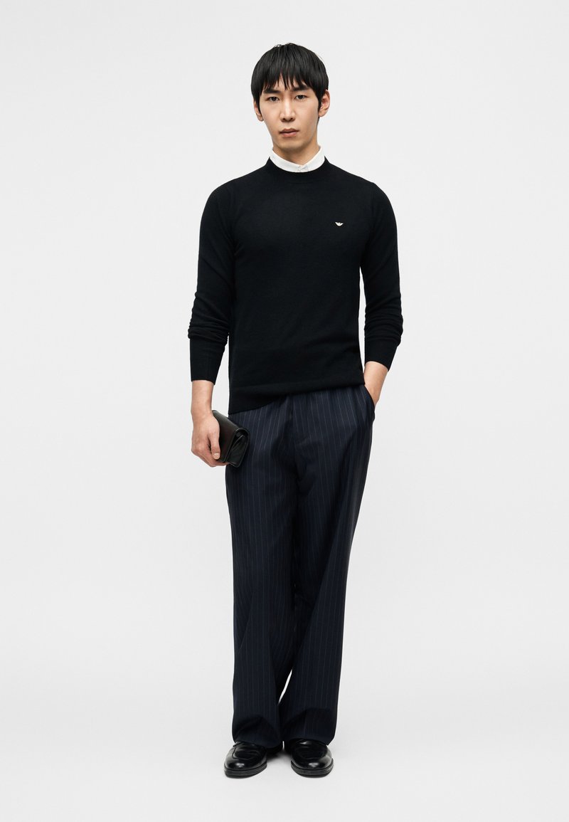 Pull noir en tricot, chemise blanche à col en dessous, pantalon bleu marine à fines rayures, pochette noire à la main, debout devant un fond uni.