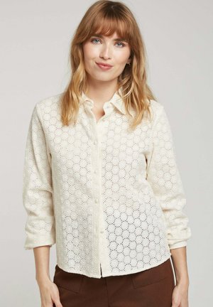 Chemise blanche à boutons, avec un motif à œillets hexagonaux, manches longues et col. Texture douce, coupe décontractée et légère brillance.