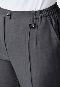 Graue Hose mit glatter Textur, ausgestattet mit einem elastischen Bund, einem Knopfverschluss und einem dekorativen Ringakzent auf der Vorderseite.