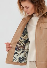 Tan leather jacket with a smooth texture, featuring a patterned inner lining with blue, cream, and brown designs. Fitted with a zip closure.