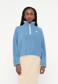 Pulover din fleece de culoare albastru deschis, cu fermoar pe un sfert, guler înalt, mâneci raglan și fermoar alb contrastant, având un mic logo pe piept.