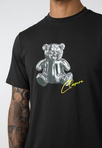 Black t-shirt featuring a shiny silver teddy bear graphic on the front, accompanied by yellow text reading "Closure" underneath.