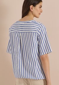 Femme aux cheveux foncés portant une chemise blanche à rayures verticales bleues et un pantalon beige, de profil sur un fond beige.