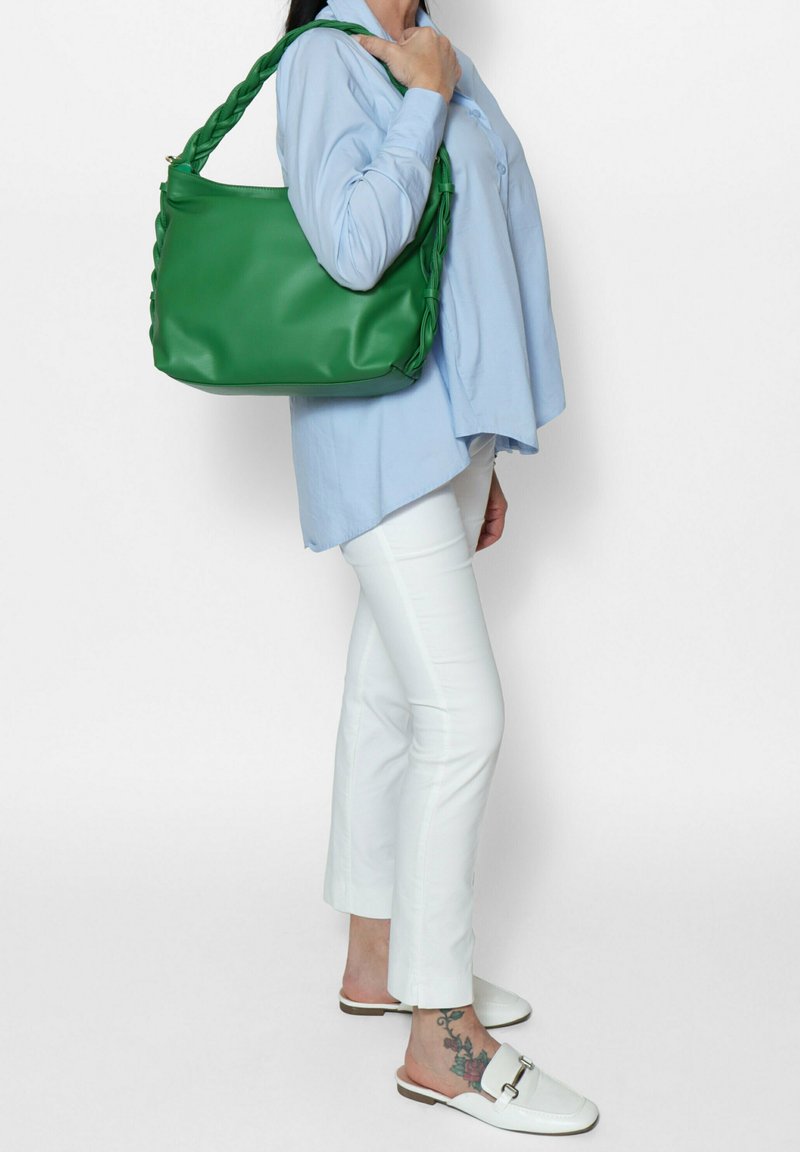 Sac à main en cuir tressé vert avec une texture douce, dotée d'une large bandoulière. Le modèle porte une chemise bleu clair et un pantalon blanc, se tenant de côté.
