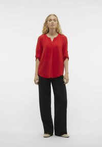Blusa rossa con scollatura a V e maniche a tre quarti, abbinata a pantaloni neri a gamba larga. Il tessuto ha una texture stropicciata.