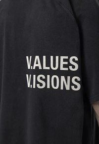 Melna kokvilnas T-kreklā ar brīvu piegriezumu. Aizmugurē ir drosmīgi balti uzraksti: "V.ALUES. V.ISIONS." Teksts ir centrēts vertikāli.