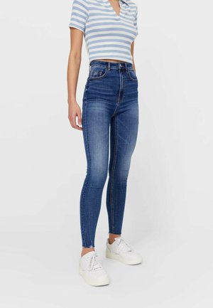 Kvinde iført en blå og hvid stribet crop top, højtaljede blå skinny jeans og hvide sneakers, stående på en hvid baggrund.