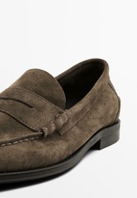 Mocassin en suède marron avec un design à couture surélevée et un petit talon noir. Présente une texture douce et des coutures visibles le long des bords.