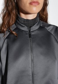 Giacca nera con zip e finitura opaca, colletto alto e maniche raglan. Indossata sopra un maglione grigio a collo alto con una texture.