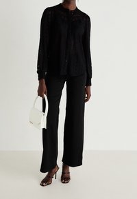 Svart spetsblus med långa ärmar och knäppning i fronten, matchad med vida svarta byxor och en liten vit handväska.