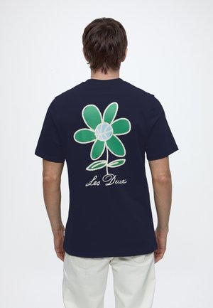 Homme portant un t-shirt bleu marine avec une grande fleur verte et un motif de basketball bleu dans le dos, étiqueté "Les Deux", associé à un pantalon blanc.
