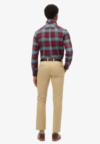 Camicia a quadri nei toni di blu, rosso e verde; pantaloni beige; cintura marrone; vista da dietro, che evidenzia il colletto e i dettagli della vestibilità della camicia.