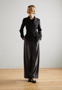 Veste noire ajustée avec une finition à boucle argentée, associée à un pantalon gris à jambes larges et des chaussures noires sur un sol en bois devant un mur clair.