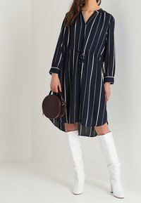 Mörkblå randig klänning med lång ärm och dragsko i midjan, matchad med glänsande vita knähöga stövlar och en rund mörkröd väska.