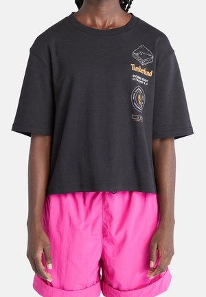 Černé oříznuté tričko s krátkými rukávy, s bílými a oranžovými motivy včetně hory a loga. Kombinováno s jasně růžovými šortkami.