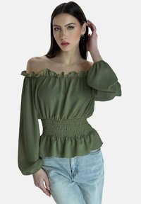 Olivgrünes Off-Shoulder-Bluse mit rüschigen Kanten, gerafftem Bund und langen Ärmeln, aus leichtem Stoff gefertigt. Kombiniert mit hellblauen Jeans.