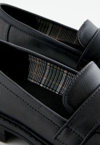 Mocassins en cuir noir avec une finition texturée et une doublure en tissu à motif écossais présentant des accents verts, blancs et oranges.