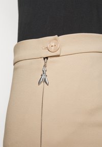 Beżowa spódnica z zamkiem błyskawicznym i srebrnym, owadopodobnym uchwytem oraz okrągłym, jasnym guzikiem w pasie. Gładka, miękka tekstura materiału.