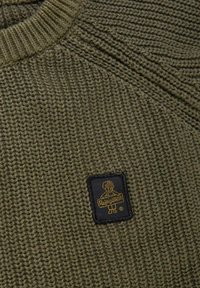 Primo piano di un maglione lavorato a maglia verde oliva con texture a costine e una patch nera che mostra un'illustrazione di un cane giallo e il testo "Resigualan".