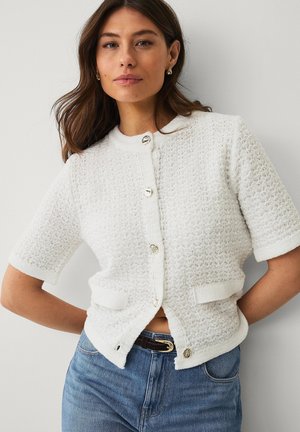 Junge Frau mit braunen Haaren, die eine strukturierte weiße kurzärmelige, geknöpfte Strickjacke und blaue Jeans trägt, steht vor einer weißen Wand.