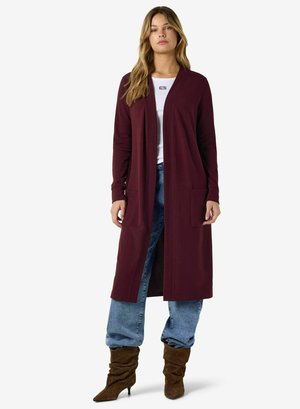 Femme portant un long cardigan bordeaux ouvert, un haut blanc, un jean bleu ample et des bottes marron à talons souples, debout devant un fond uni.