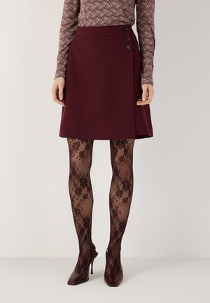 Frau trägt einen burgunderfarbenen A-Linien-Rock mit drei Knöpfen, schwarze florale Spitzenstrumpfhosen, spitze burgunderfarbene High Heels und ein gemustertes langärmeliges Oberteil.