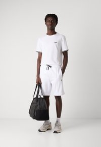 T-shirt blanc à manches courtes avec des accents noirs et un logo, shorts blancs à cordon, baskets grises et un sac fourre-tout noir. Design épuré, look décontracté.