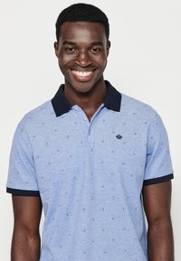 Ljusblå polotröja med marinblå krage och ärmslut, med ett upprepande mönster av små blommor och broderad logotyp på bröstet.