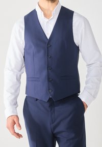 Námořnický modrý vest s přiléhavým střihem, včetně šesti knoflíků, dvou předních kapes a hladké textury látky. Nosený přes bílou košili.
