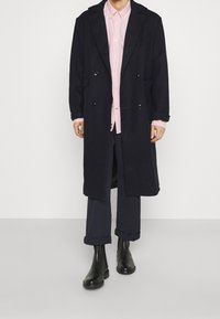 Cappotto doppiopetto blu navy realizzato in tessuto spesso, con una silhouette lunga, completato da una camicia rosa chiaro sotto e pantaloni scuri.