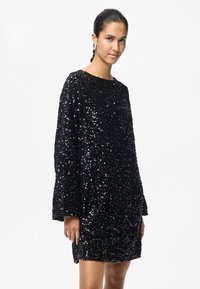 Kobieta z ciemnymi włosami związanymi w koński ogon, ubrana w długą, czarną sukienkę z cekinami, stoi na tle gładkiego białego tła.