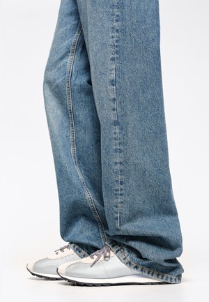 Jeans de denim azul claro con un corte relajado, combinados con zapatillas deportivas plateadas que tienen acentos gris claro y una suela texturizada.