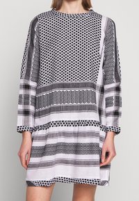 Vestido de manga larga en blanco y negro. Presenta patrones geométricos, variaciones en el grosor de las rayas y un corte relajado con un dobladillo fruncido.
