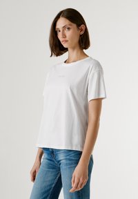 Λευκό βαμβακερό T-shirt με κοντά μανίκια, στρογγυλή λαιμόκοψη και διακριτικό λογότυπο στο κέντρο. Φοριέται με εφαρμοστά μπλε τζιν.