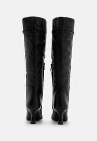 Botas de cuero negras hasta la rodilla con un diseño ajustado, que cuentan con tacones curvados y cremalleras en los lados exteriores para facilitar su uso.