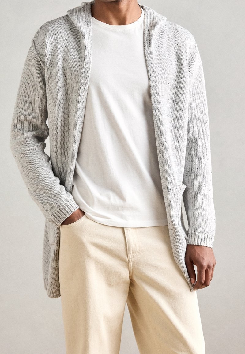 Homme portant un cardigan à capuche tricoté gris clair, un t-shirt blanc et un pantalon beige, une main dans la poche, sur fond neutre.
