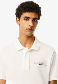 Polo bianco con colletto, maniche corte e logo "LACOSTE" ricamato con accentuazione verde. Tessuto testurizzato con vestibilità classica.