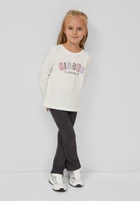 Junges Mädchen mit blonden Haaren, das ein weißes Langarmshirt mit dem Text "Der Zirkus ist um mich herum", dunkle Hose und weiße Sneakers trägt, steht vor einer grauen Wand.